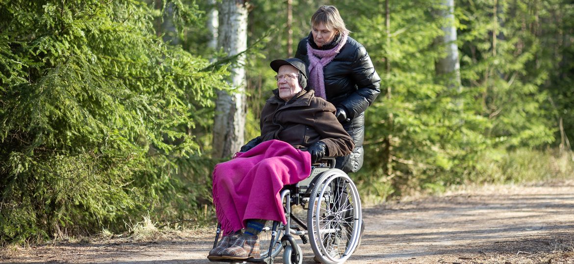 Valokuva yrityksestä Dementiakoti Villa Tapiola: hoitaja ulkoilemassa lähimetsässä pyörätuolissa olevan asukkaan kanssa